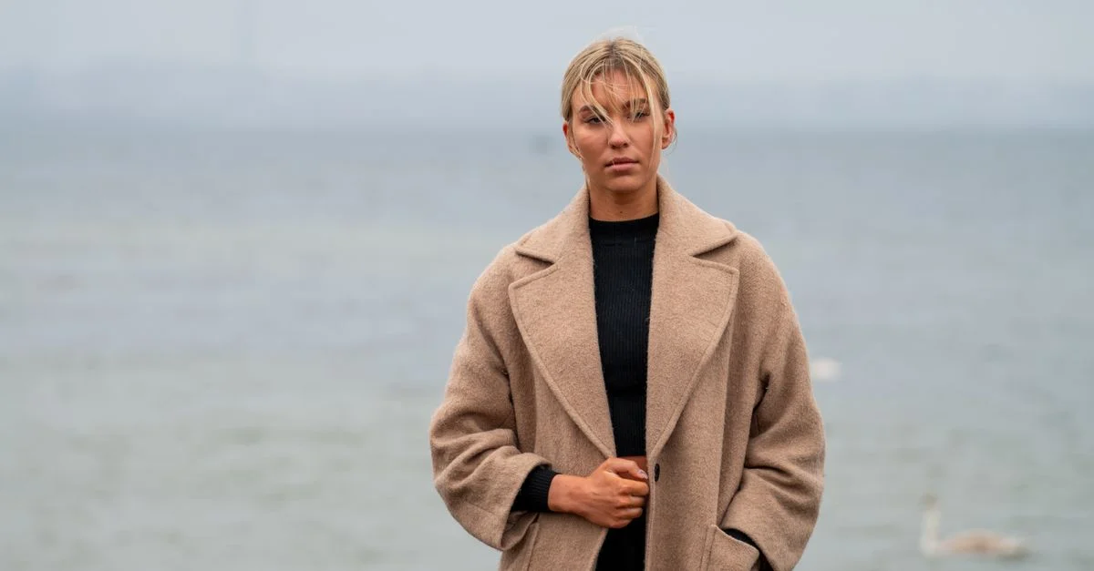 Une femme stylée en manteau beige au bord de la baie de Tallinn, dégageant élégance moderne et sérénité.